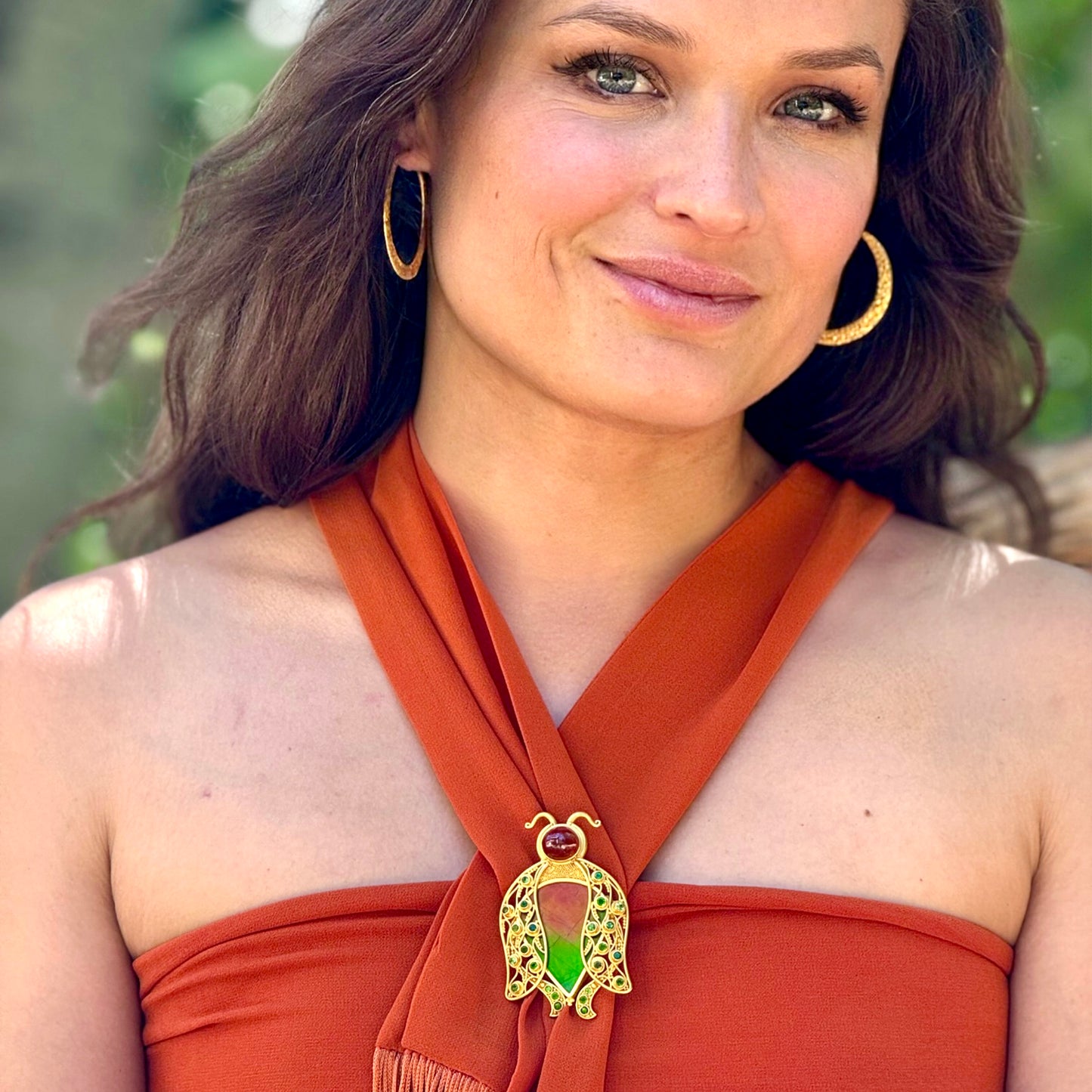 Woman wearing a rust-colored top with a decorative brooch, outdoors.