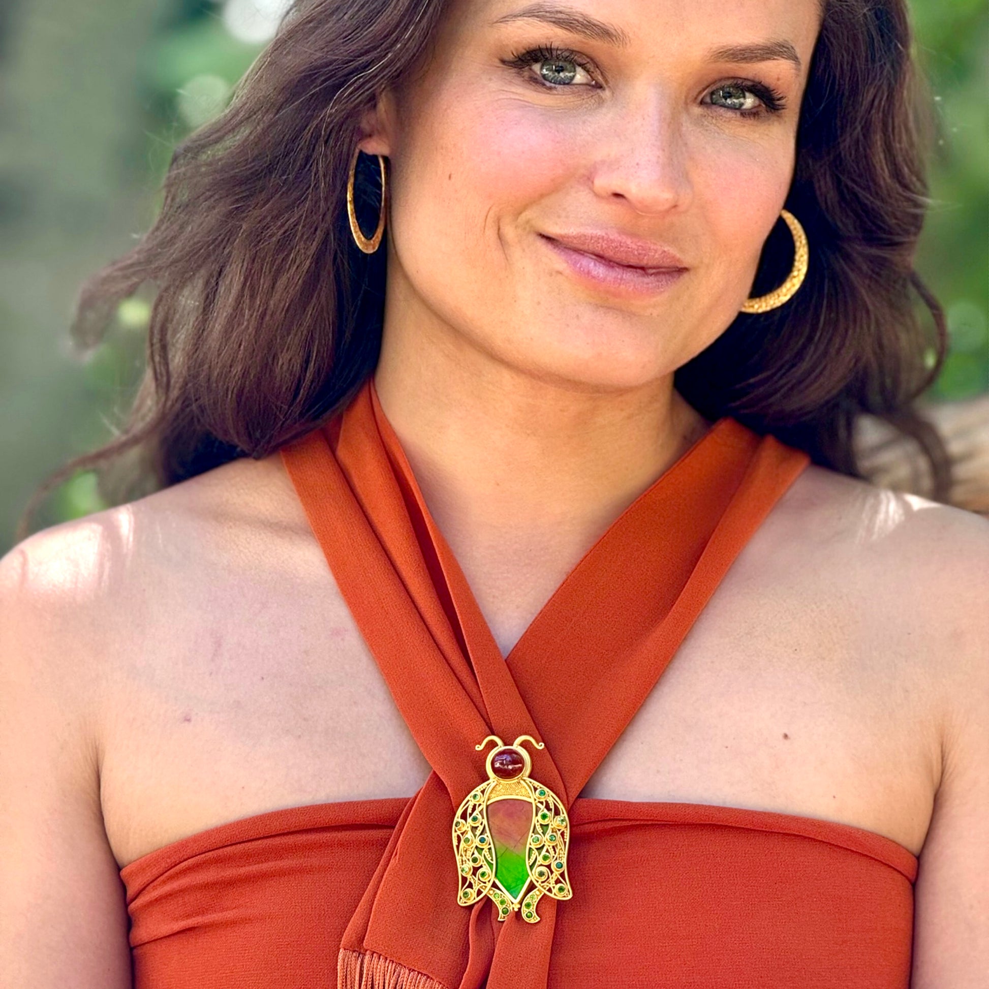 Woman wearing a rust-colored top with a decorative brooch, outdoors.
