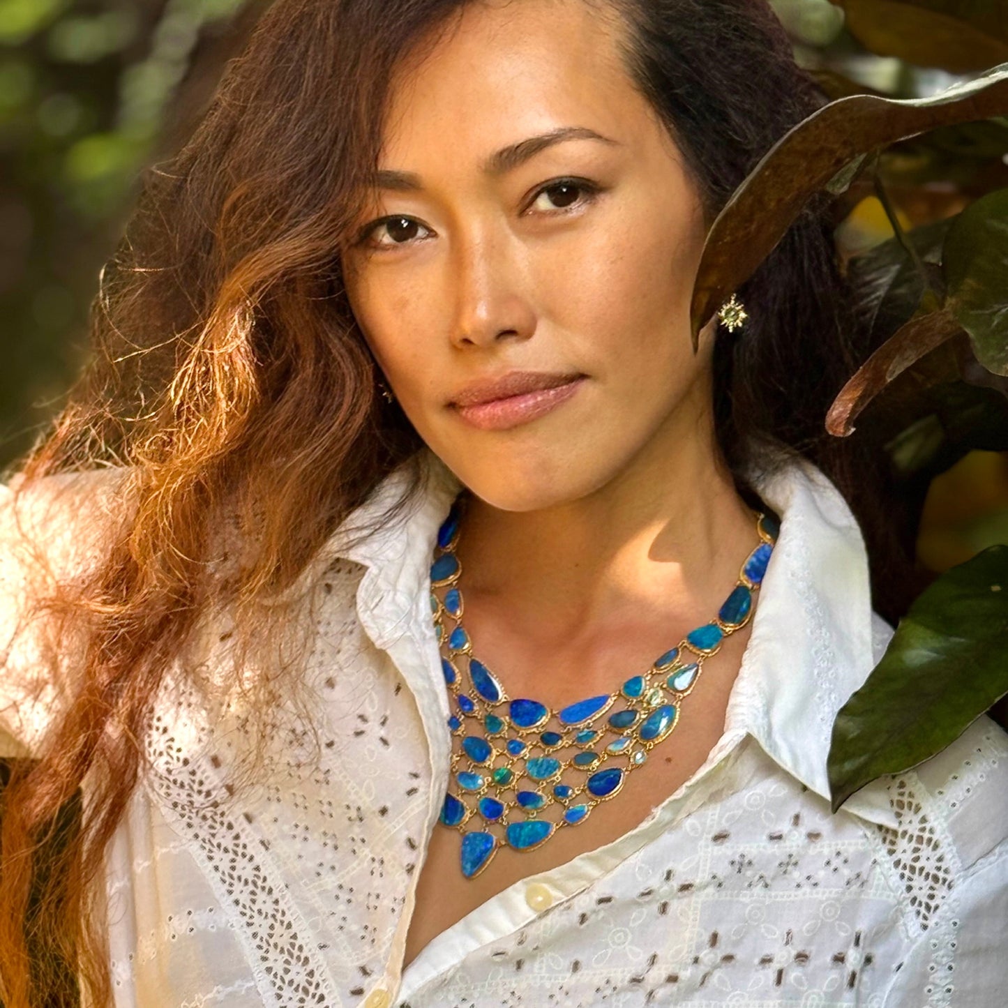 Woman wearing a white blouse and blue necklace with greenery in the background