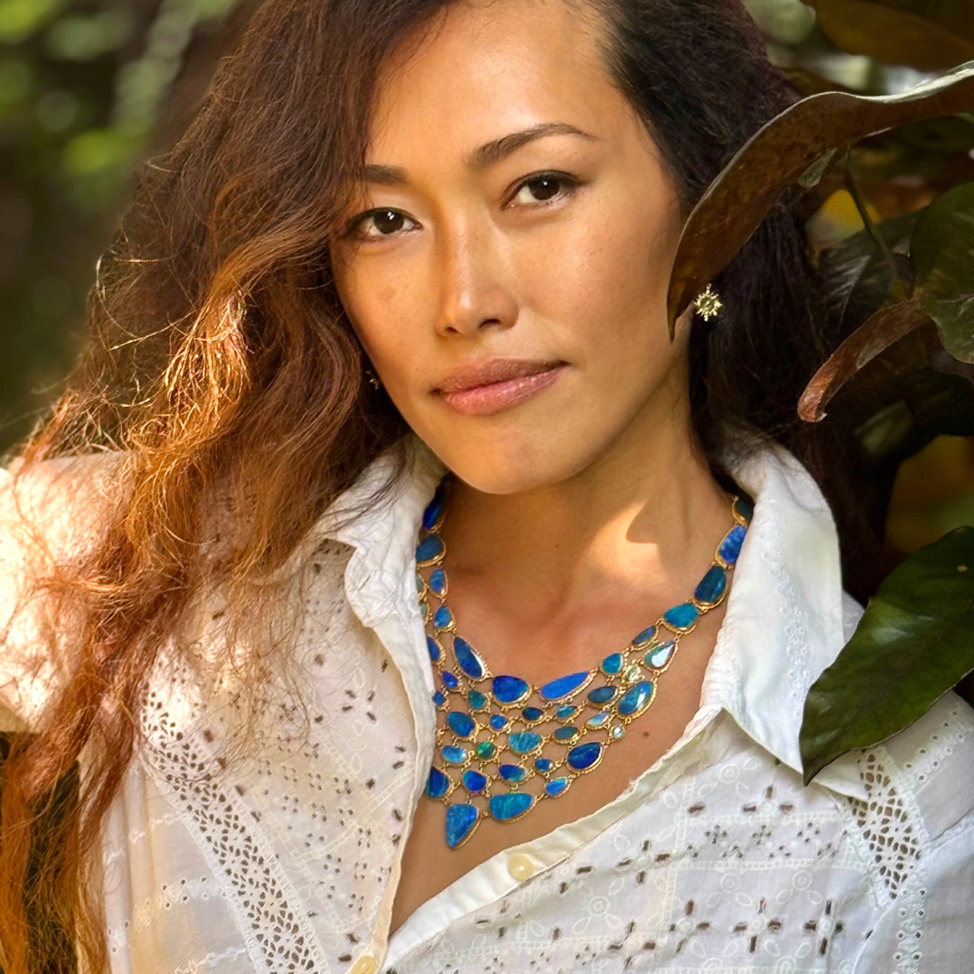 Woman wearing a white blouse and blue necklace with greenery in the background