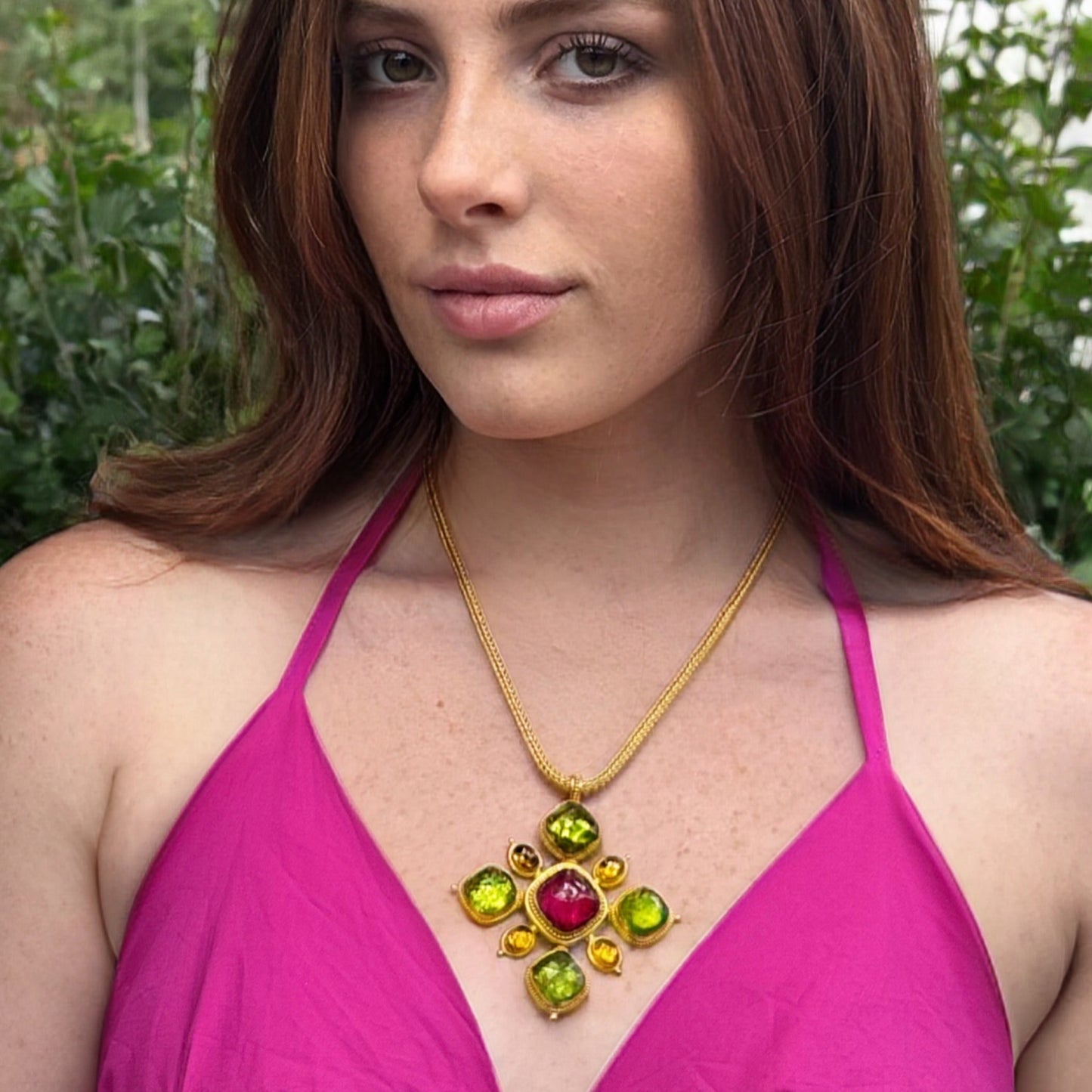 Woman wearing a pink top and colorful necklace with a green and red gemstone, standing outdoors.