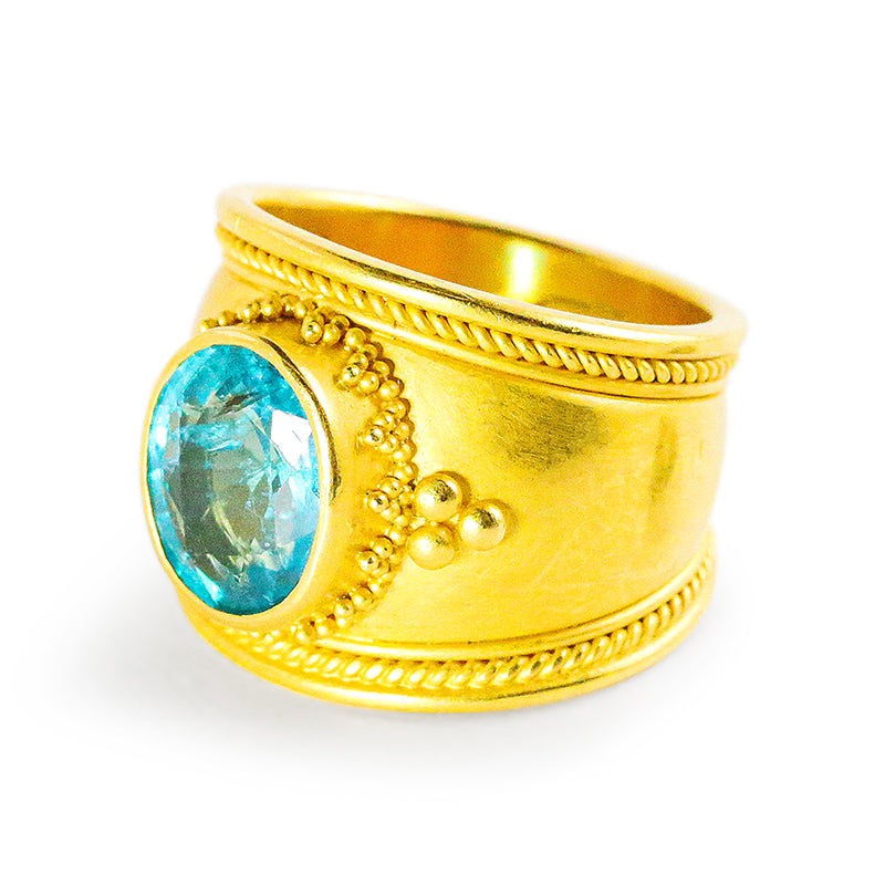 A gold-colored band ring with a large Caribbean-blue faceted oval apatite in the center, featuring granulation detail on the band.
